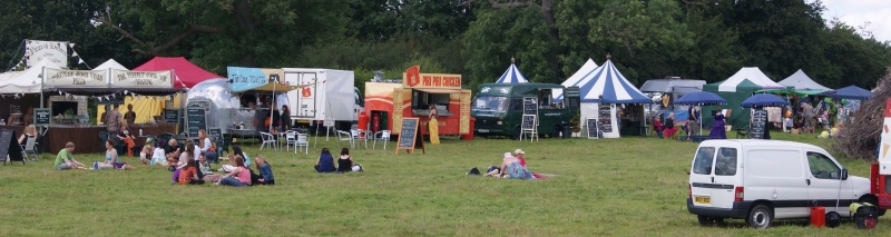 A line of catering vans, trailers, stalls and tents, with tables, chairs and customers in front of them.